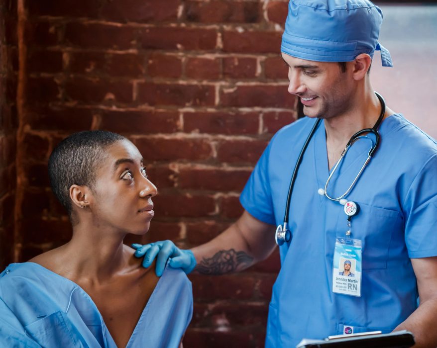 A doctor in blue scrubs interacting with a patient in a hospital setting, with a brick wall in the background