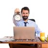 man in a shirt and tie sits at a desk with a laptop and a clock, focused on his work