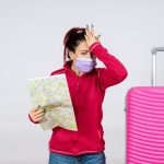 A woman in a mask holds a map while standing next to a pink suitcase, ready for her journey