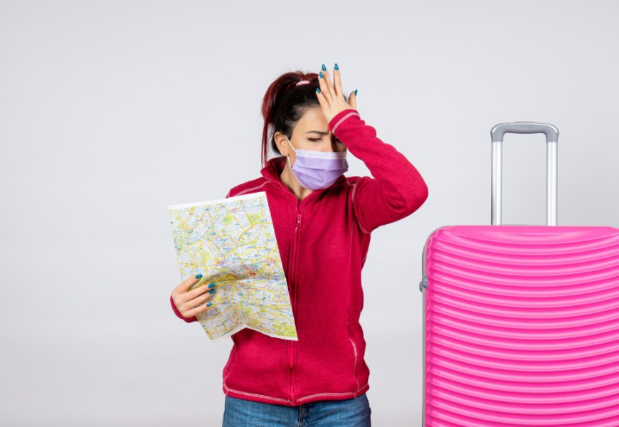 A woman in a mask holds a map while standing next to a pink suitcase, ready for her journey