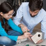 man and a girl collaborate on building a birdhouse, surrounded by tools and wood materials